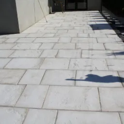 Jasna, prostokątna nawierzchnia z kostki, z ciemniejszym obrzeżem. Widoczne cienie drzew i fragment budynku w tle. Ujęcie z niskiej perspektywy, podkreślające regularność wzoru.