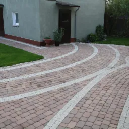 Efektowna nawierzchnia z kostki brukowej w odcieniach brązu, z białymi pasami dekoracyjnymi, tworząca wzór łuków. Widok na dom i zadbany trawnik.