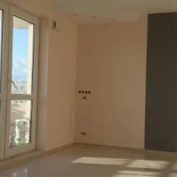 Jasne wnętrze po remoncie: ściany w odcieniach beżu i szarości, okna z widokiem na miasto, widoczne puszki z farbą, kaloryfer i instalacja elektryczna.