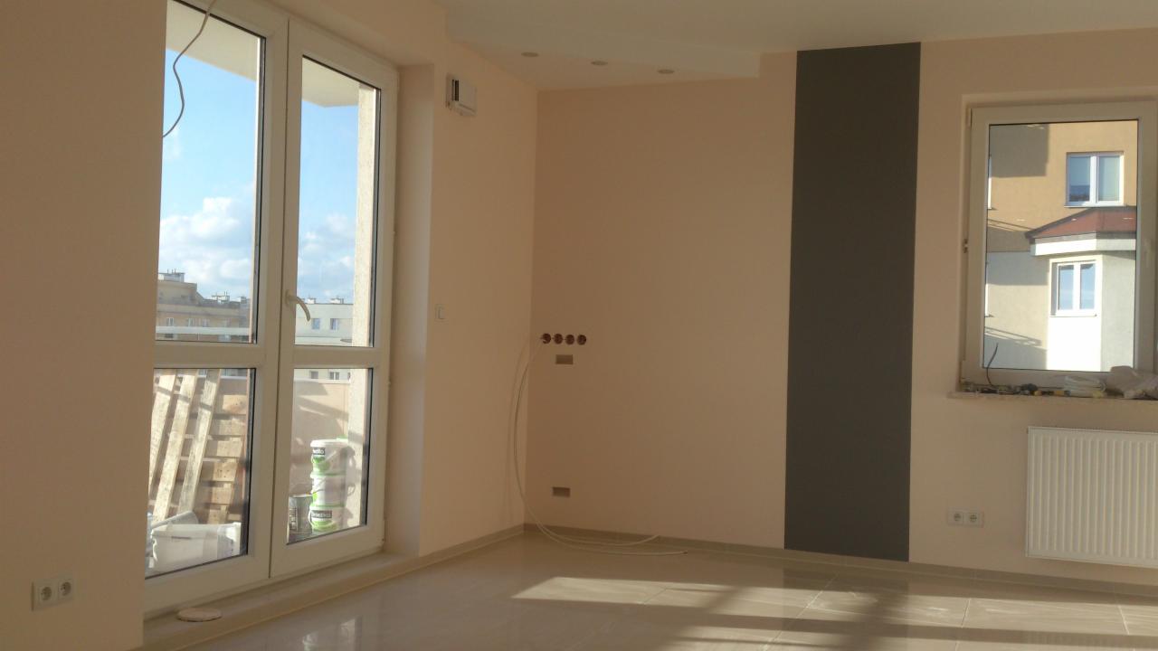 Jasne wnętrze po remoncie: ściany w odcieniach beżu i szarości, okna z widokiem na miasto, widoczne puszki z farbą, kaloryfer i instalacja elektryczna.