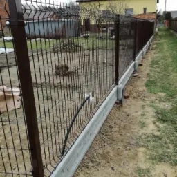 Nowo zamontowana siatka ogrodzeniowa z brązowymi słupkami i betonową podmurówką, widoczne ślady prac ziemnych, w tle żółty dom z ceglanym kominem.