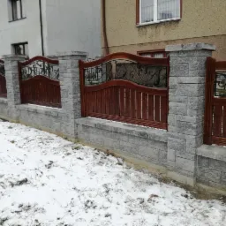 Nowoczesne ogrodzenie z drewnianymi przęsłami i elementami kutymi, osadzone na niskim murku z szarej kostki, pokryte częściowo śniegiem.