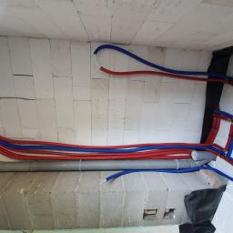 AB Complex - Wnętrze budynku w trakcie budowy, widoczne rury instalacyjne w kolorach czerwonym i niebieskim prowadzone wzdłuż ściany z betonu komórkowego, oraz szara rura kanalizacyjna, na ścianie widoczne...