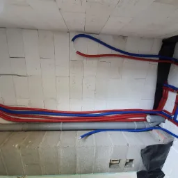 Wnętrze budynku w trakcie budowy, widoczne rury instalacyjne w kolorach czerwonym i niebieskim prowadzone wzdłuż ściany z betonu komórkowego, oraz szara rura kanalizacyjna, na ścianie widoczne...