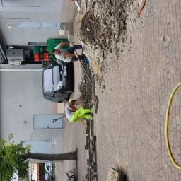 AB Complex - Dwóch robotników w trakcie prac ziemnych przy układaniu kostki brukowej na chodniku, z widocznymi narzędziami, stertą piasku i częściowo ułożoną nawierzchnią, na tle budynku z zaparkowanym samochodem.