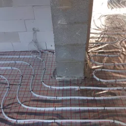 Rozłożone białe rurki na folii z czerwoną siatką, mocowane czarnymi klipsami, widok na surowy betonowy słup i fragment ściany z bloczk&oacute;w.