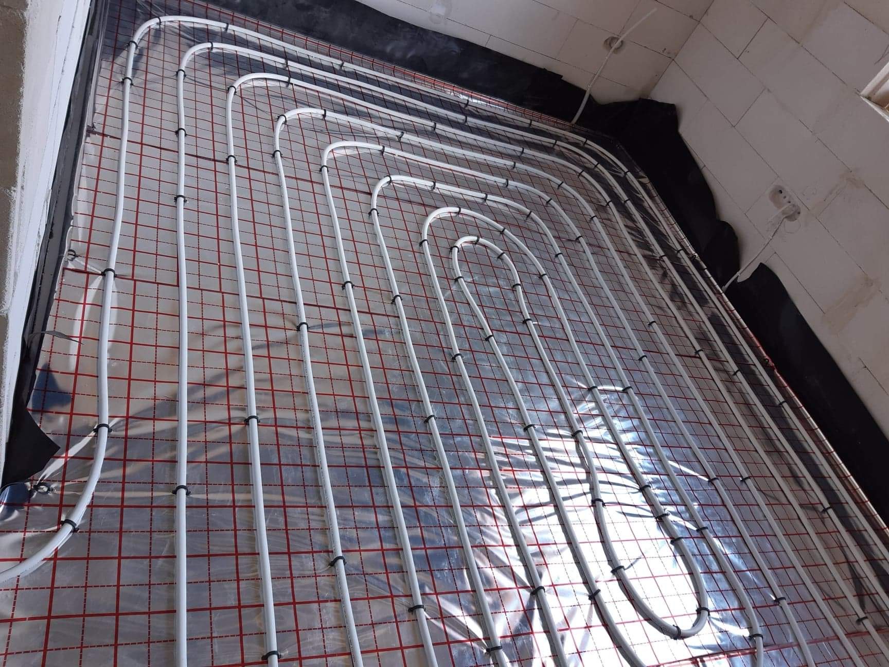 Biała: Rozłożone białe rury ogrzewania podłogowego na siatce z czerwonymi kwadratami, czarna folia izolacyjna przy ścianach, surowy stan wykończenia.