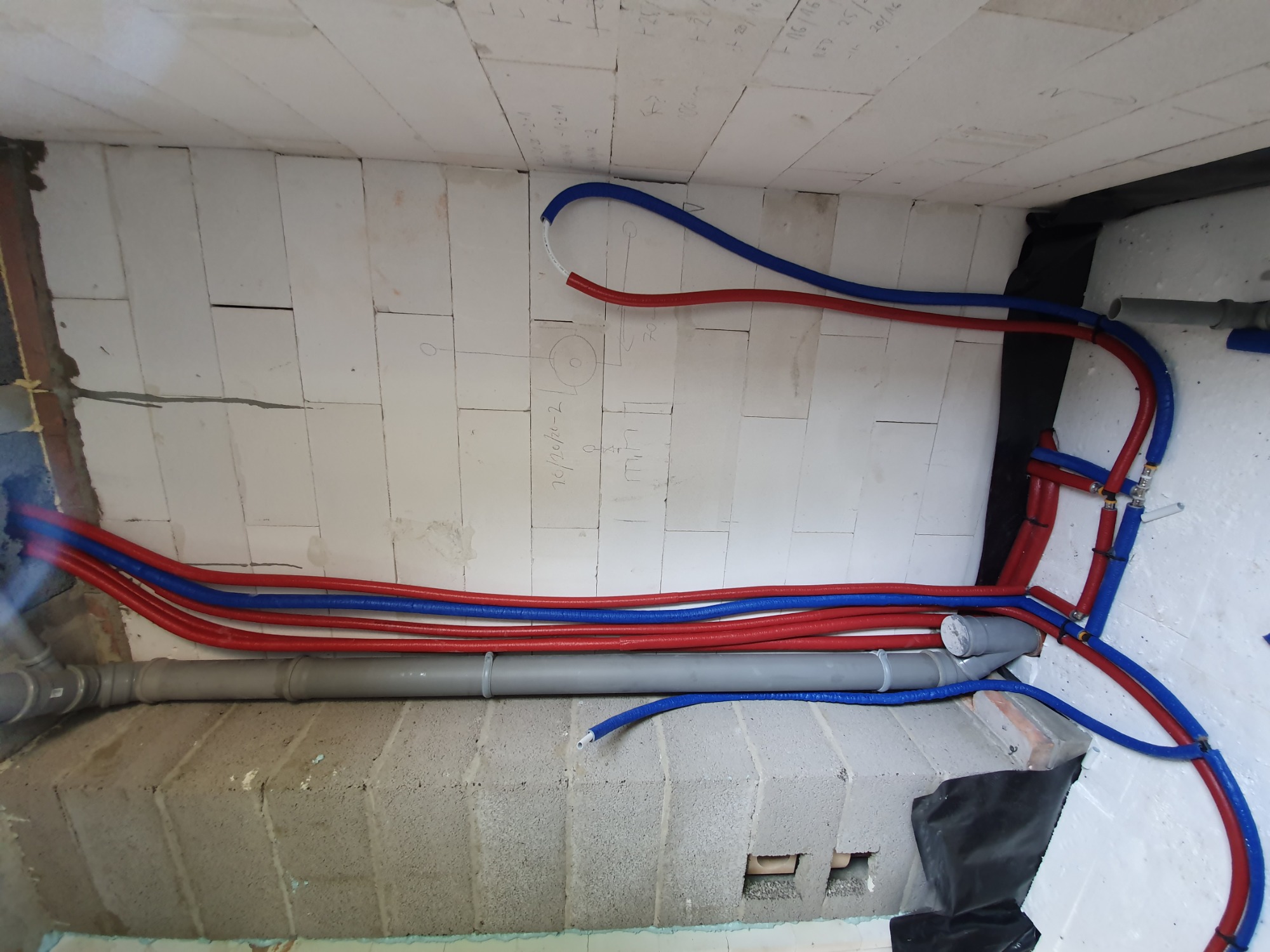 Wnętrze budynku w trakcie budowy, widoczne rury instalacyjne w kolorach czerwonym i niebieskim prowadzone wzdłuż ściany z betonu komórkowego, oraz szara rura kanalizacyjna, na ścianie widoczne...