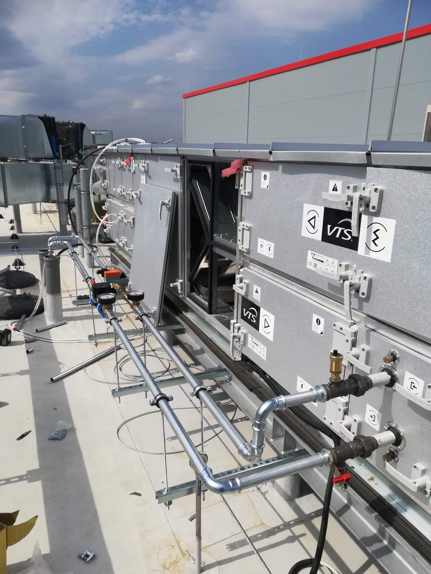 Szara centrala wentylacyjna VTS na dachu budynku komercyjnego z widocznymi rurami, manometrami i otwartym panelem dostępowym.