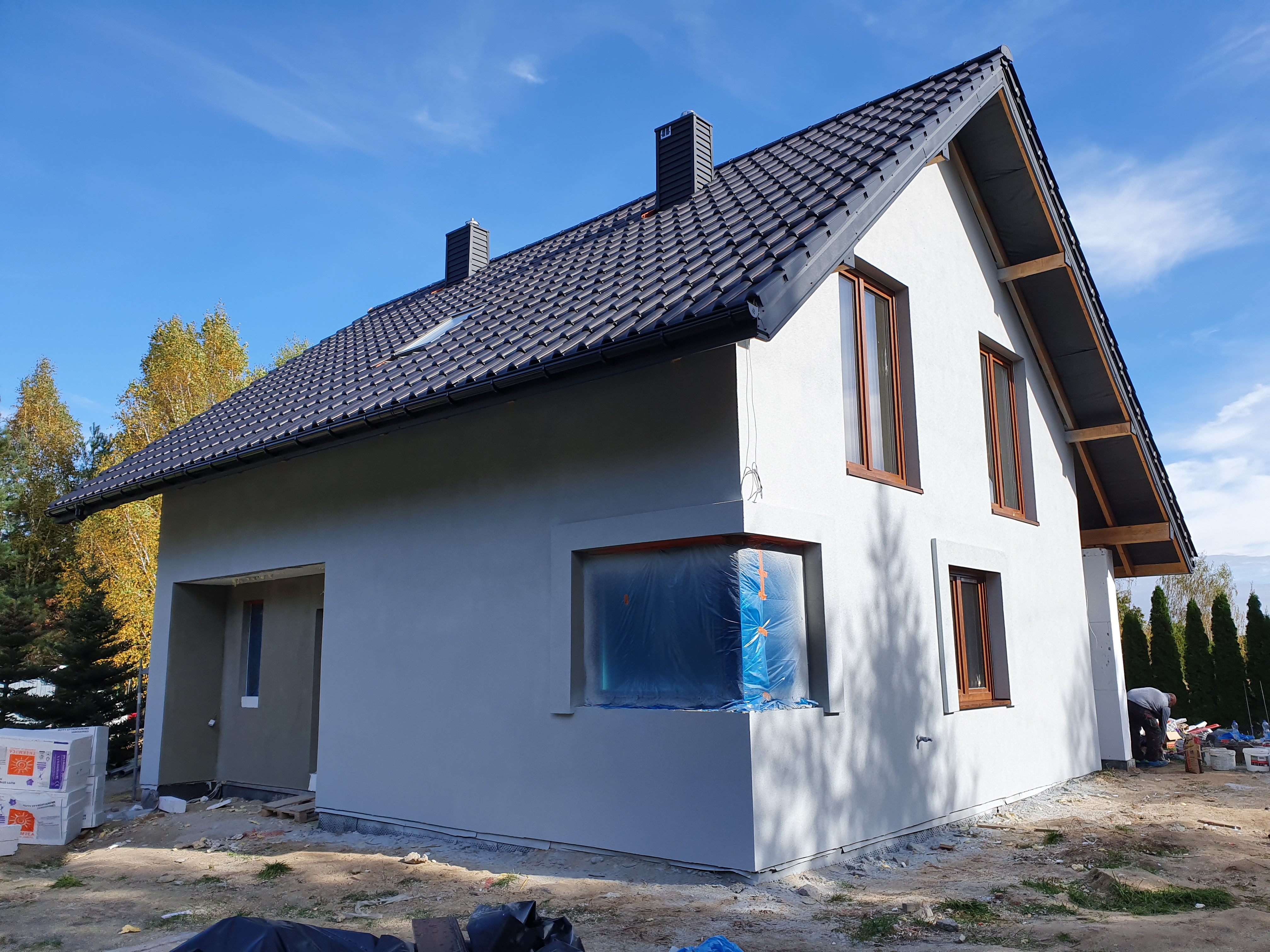Nowoczesny dom jednorodzinny z grafitową dachówką i szarym tynkiem zewnętrznym w trakcie budowy. Okna zabezpieczone folią. Widoczne materiały budowlane.