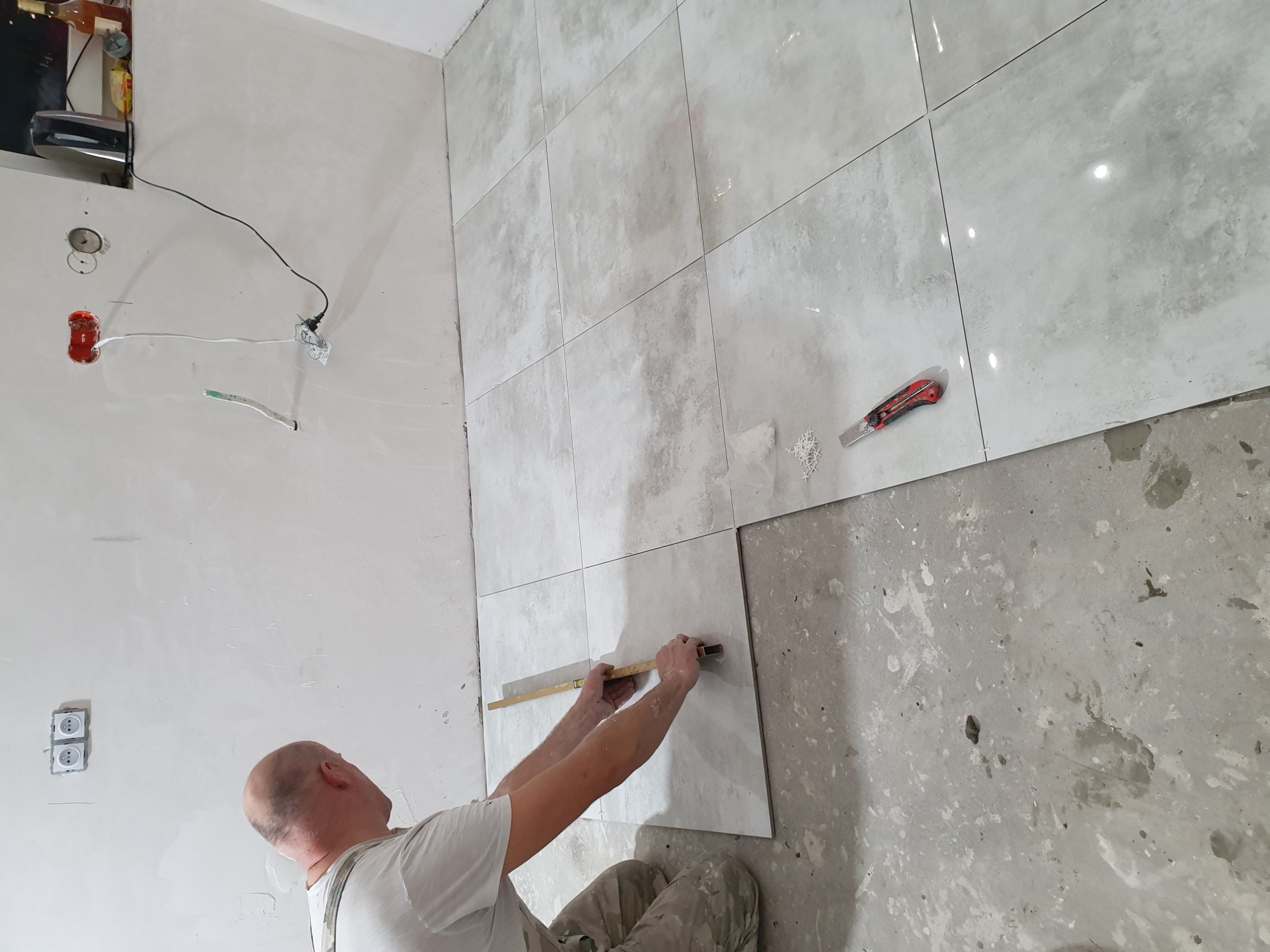 Układanie jasnych, kwadratowych płytek ściennych z użyciem poziomicy, widoczny fragment podłogi w trakcie prac wykończeniowych i gniazdka elektryczne bez osprzętu