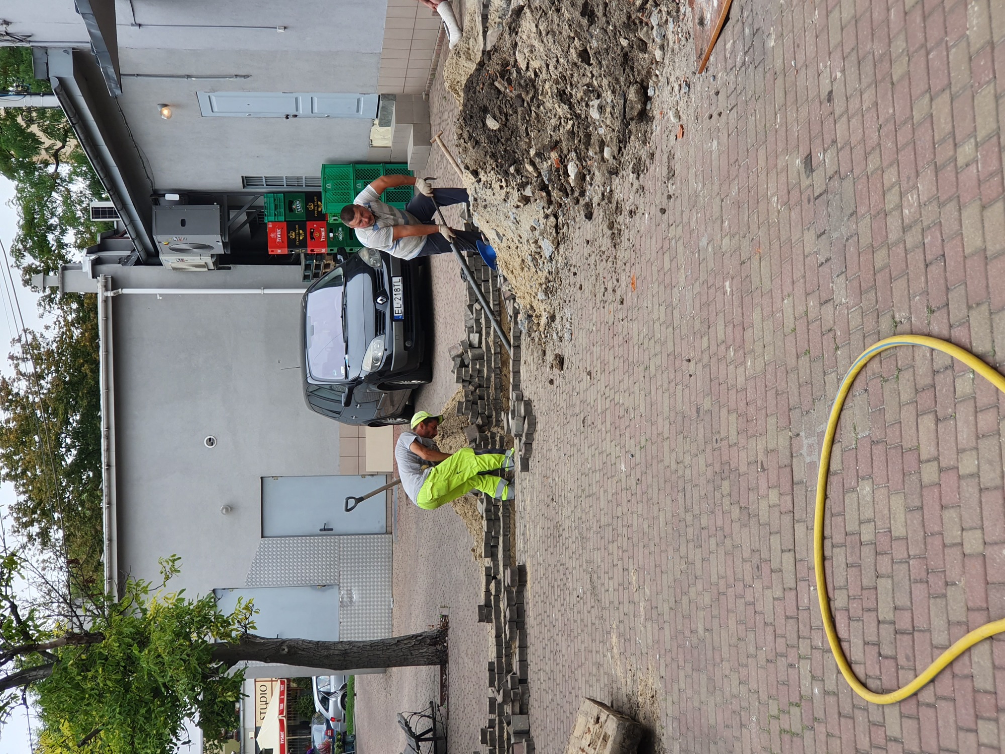 Dwóch robotników w trakcie prac ziemnych przy układaniu kostki brukowej na chodniku, z widocznymi narzędziami, stertą piasku i częściowo ułożoną nawierzchnią, na tle budynku z zaparkowanym samochodem.
