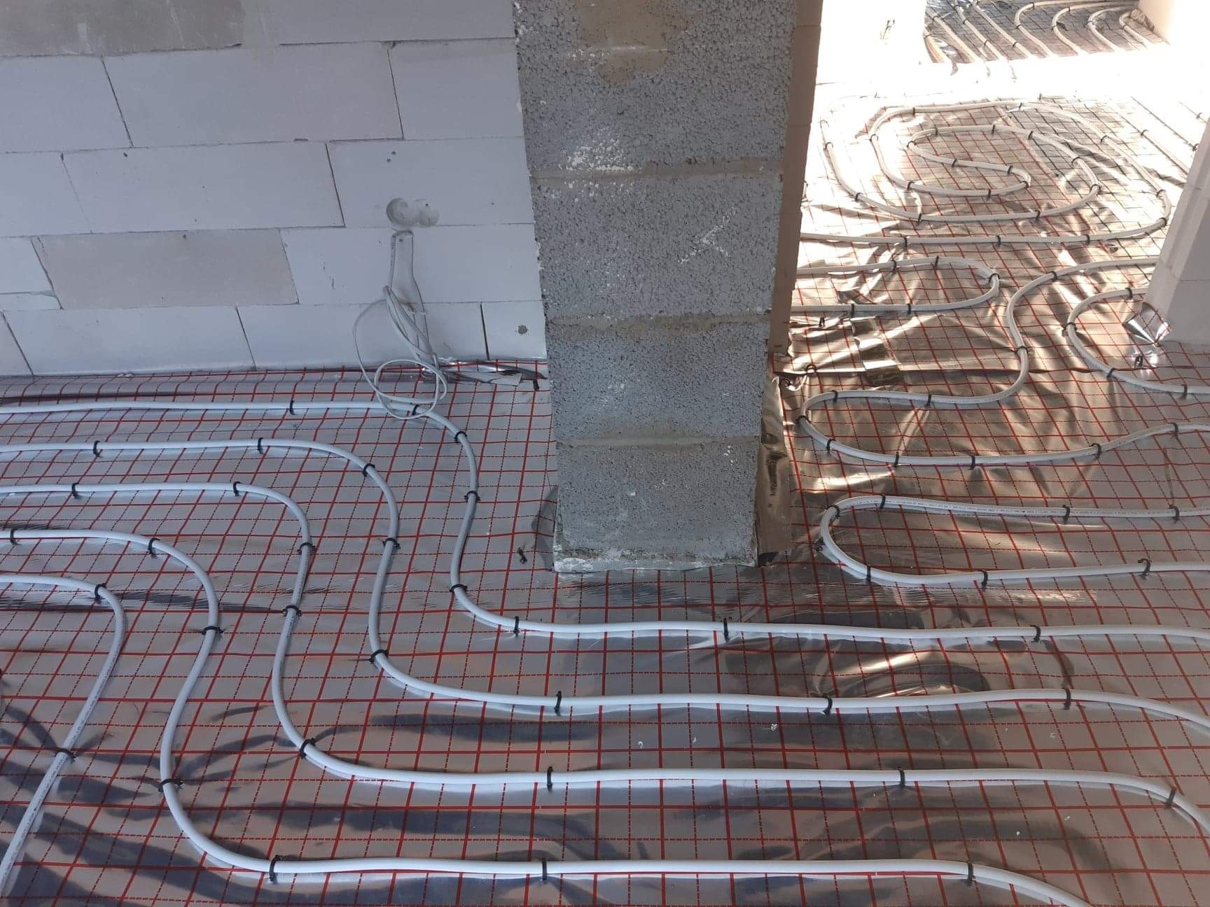 Rozłożone białe rurki na folii z czerwoną siatką, mocowane czarnymi klipsami, widok na surowy betonowy słup i fragment ściany z bloczków.