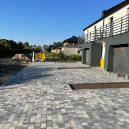 Nowo ułożona, szara kostka brukowa prowadząca do nowoczesnego domu z dwoma garażami, z widocznym przygotowanym miejscem pod trawnik i stertą piasku w tle.