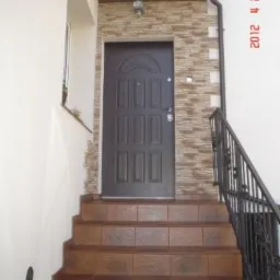 Wejście do domu z brązowymi schodami wyłożonymi płytkami, ciemnymi drzwiami i elewacją z kamienia dekoracyjnego w odcieniach beżu i brązu, widoczne rury spustowe.