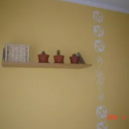 Żółta ściana z dekoracyjnym białym wzorem, na której wisi drewniana półka z trzema kaktusami w terakotowych doniczkach i wiklinowym koszykiem.