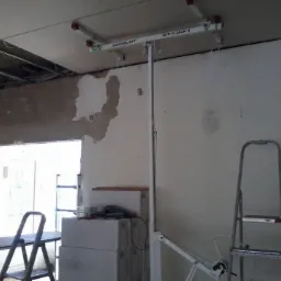 Montaż płyty gipsowo-kartonowej na suficie za pomocą podnośnika Goldblatt GYP-C-JACK II, widoczne niedokończone ściany i drabiny.