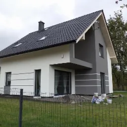 Nowoczesny dom jednorodzinny z dwukolorową elewacją, szarą strefą z balkonem i białymi ścianami, widoczne puszki z farbą i materiały budowlane przy ogrodzeniu.