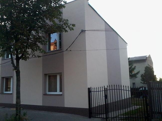 Dwukondygnacyjny budynek po termomodernizacji z elewacją w odcieniach szarości, widoczny zza czarnego, metalowego ogrodzenia i drzewa.