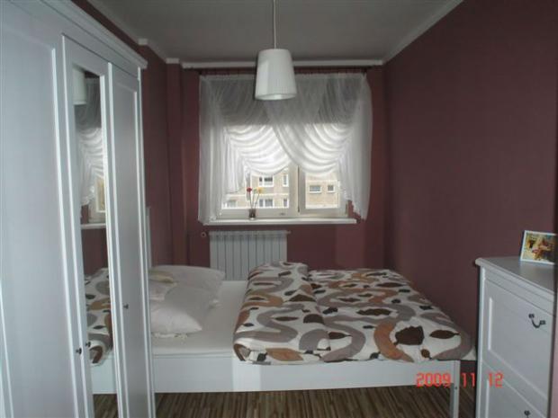 Pokój z łóżkiem, szafą z lustrem i oknem z firankami. Ściany pomalowane na kolor śliwkowy.