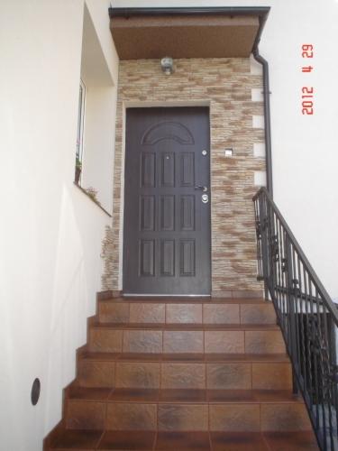 Wejście do domu z brązowymi schodami wyłożonymi płytkami, ciemnymi drzwiami i elewacją z kamienia dekoracyjnego w odcieniach beżu i brązu, widoczne rury spustowe.