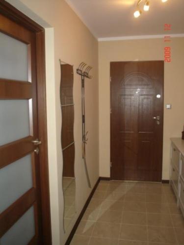 Przedpokój z drewnianymi drzwiami z przeszkleniami, lustrem o falistym kształcie, wieszakiem na ubrania i brązowymi płytkami podłogowymi.