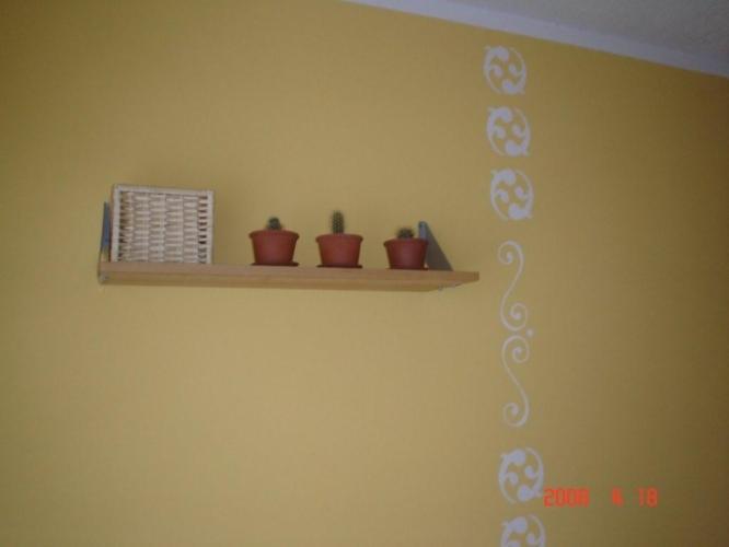 Żółta ściana z dekoracyjnym białym wzorem, na której wisi drewniana półka z trzema kaktusami w terakotowych doniczkach i wiklinowym koszykiem.