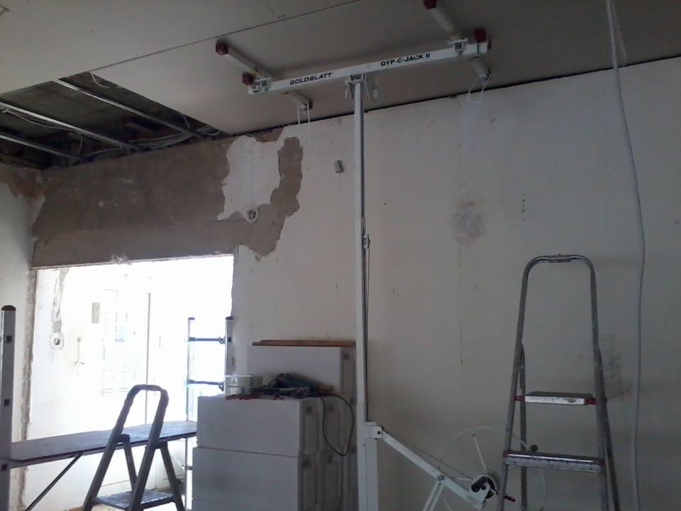 Montaż płyty gipsowo-kartonowej na suficie za pomocą podnośnika Goldblatt GYP-C-JACK II, widoczne niedokończone ściany i drabiny.