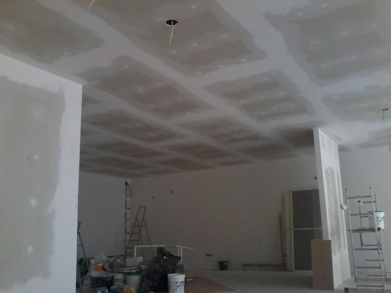 Montaż płyt gipsowo-kartonowych na suficie, widoczne łączenia i otwory na oświetlenie, pomieszczenie w trakcie wykańczania, drabina i materiały budowlane w tle.