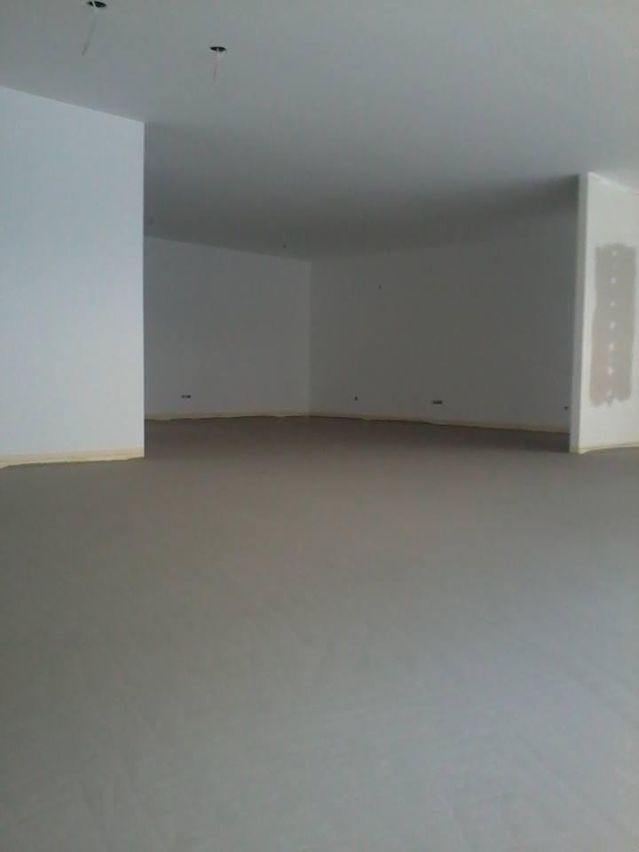 Surowe wnętrze z szarą wylewką podłogową, białe ściany przygotowane do dalszych prac wykończeniowych, widoczne kable wystające z sufitu.