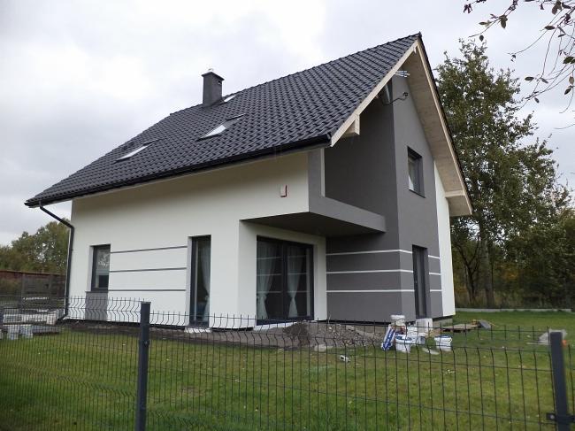 Nowoczesny dom jednorodzinny z dwukolorową elewacją, szarą strefą z balkonem i białymi ścianami, widoczne puszki z farbą i materiały budowlane przy ogrodzeniu.