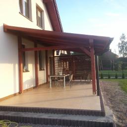 Zakład Stolarski - Zadaszony taras z drewnianą konstrukcją przy domu, wyłożony jasnymi płytkami. Widoczne meble ogrodowe i elementy dekoracyjne. Ujęcie w słoneczny dzień.