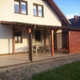 Zakład Stolarski - Zadaszony taras z drewnianą konstrukcją i ażurową ścianką, stół z krzesłami. Jasne płytki na podłodze, obrzeże z kostki. Dom z beżową elewacją w tle, błękitne niebo.