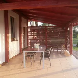 Zadaszony taras z drewnianą pergolą i ażurowymi ściankami. Na jasnej terakocie stoi stół z krzesłami. Całość oświetlona popołudniowym słońcem.