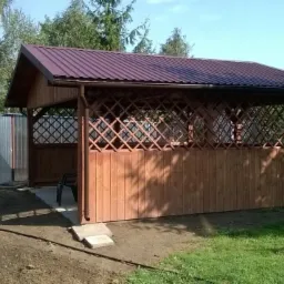 Drewniana altana z brązowym dachem i ażurowymi ścianami, otoczona zielenią. Widoczny fragment ogrodu i metalowe elementy konstrukcyjne z boku.