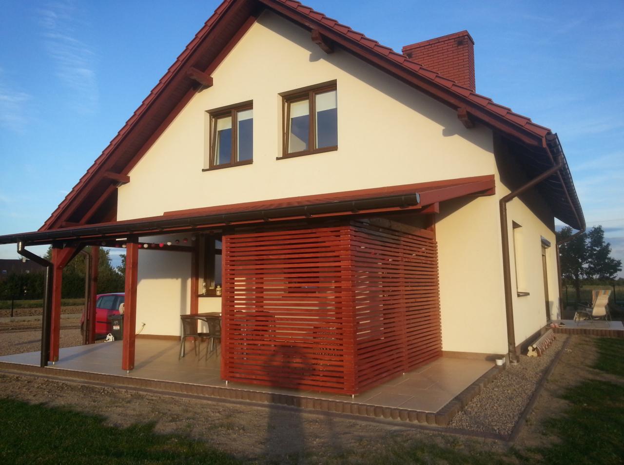 Drewniana pergola z ażurowymi ścianami, przylegająca do domu z czerwonym dachem. Na tarasie stolik i krzesła. Jasne tło nieba i trawnika.
