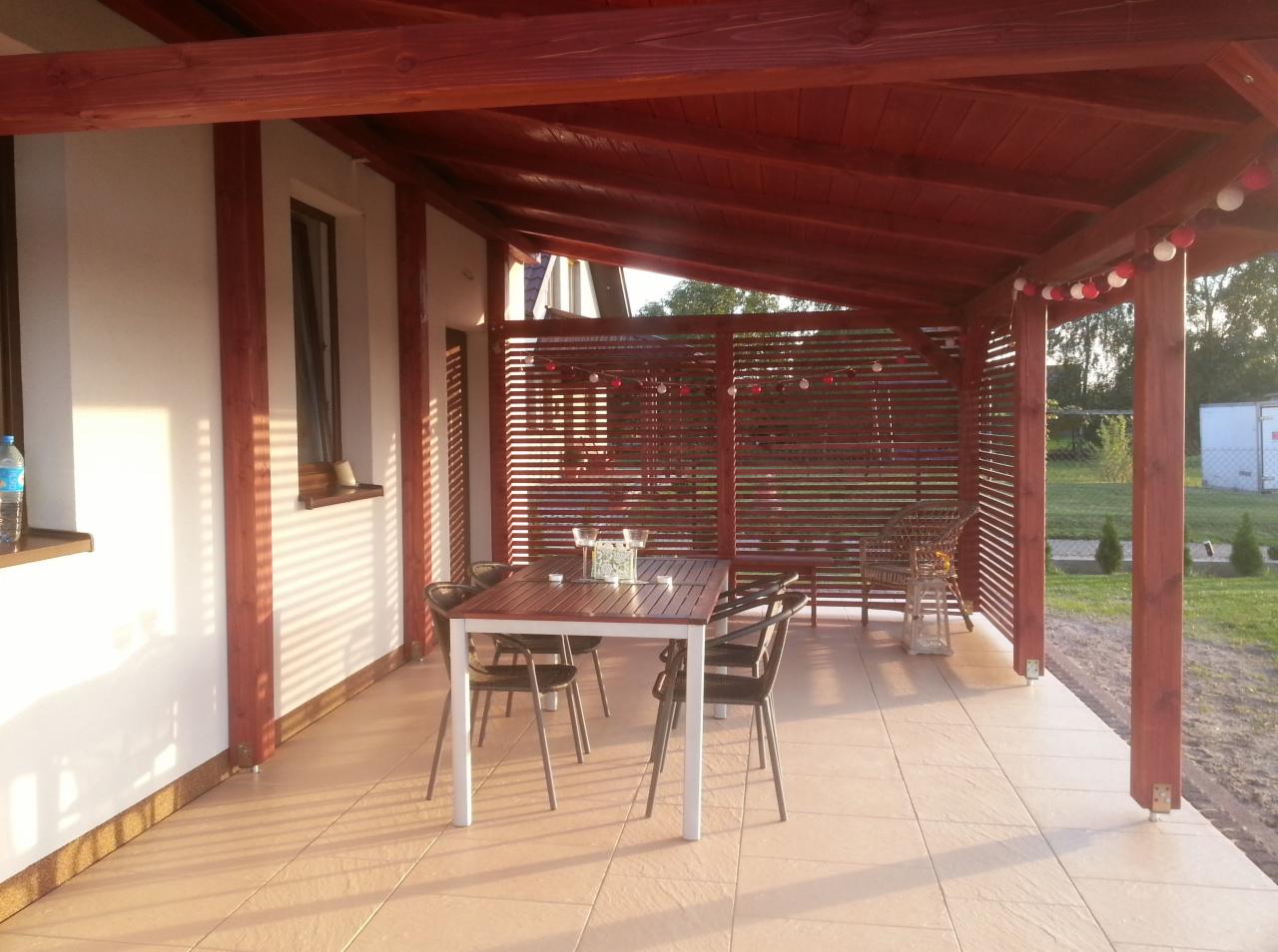 Zadaszony taras z drewnianą pergolą i ażurowymi ściankami. Na jasnej terakocie stoi stół z krzesłami. Całość oświetlona popołudniowym słońcem.