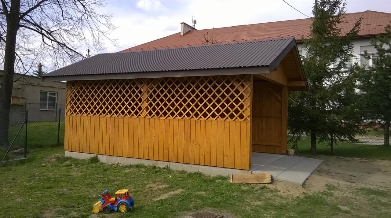 Drewniana konstrukcja z kratownicą i ciemnym dachem na betonowej podstawie, otoczona zielenią. W tle budynek. Na trawie leży zabawka.