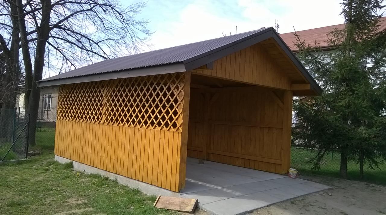 Żółta, drewniana konstrukcja z kratownicą, prawdopodobnie wiata lub altana, z szarym dachem, na betonowej podstawie. W tle drzewa i budynek.