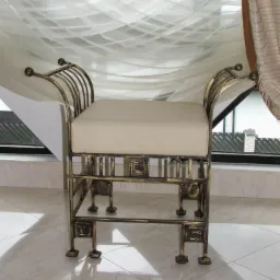 Metaloplastyka: Kute siedzisko z beżową poduszką na tle okna z firankami.