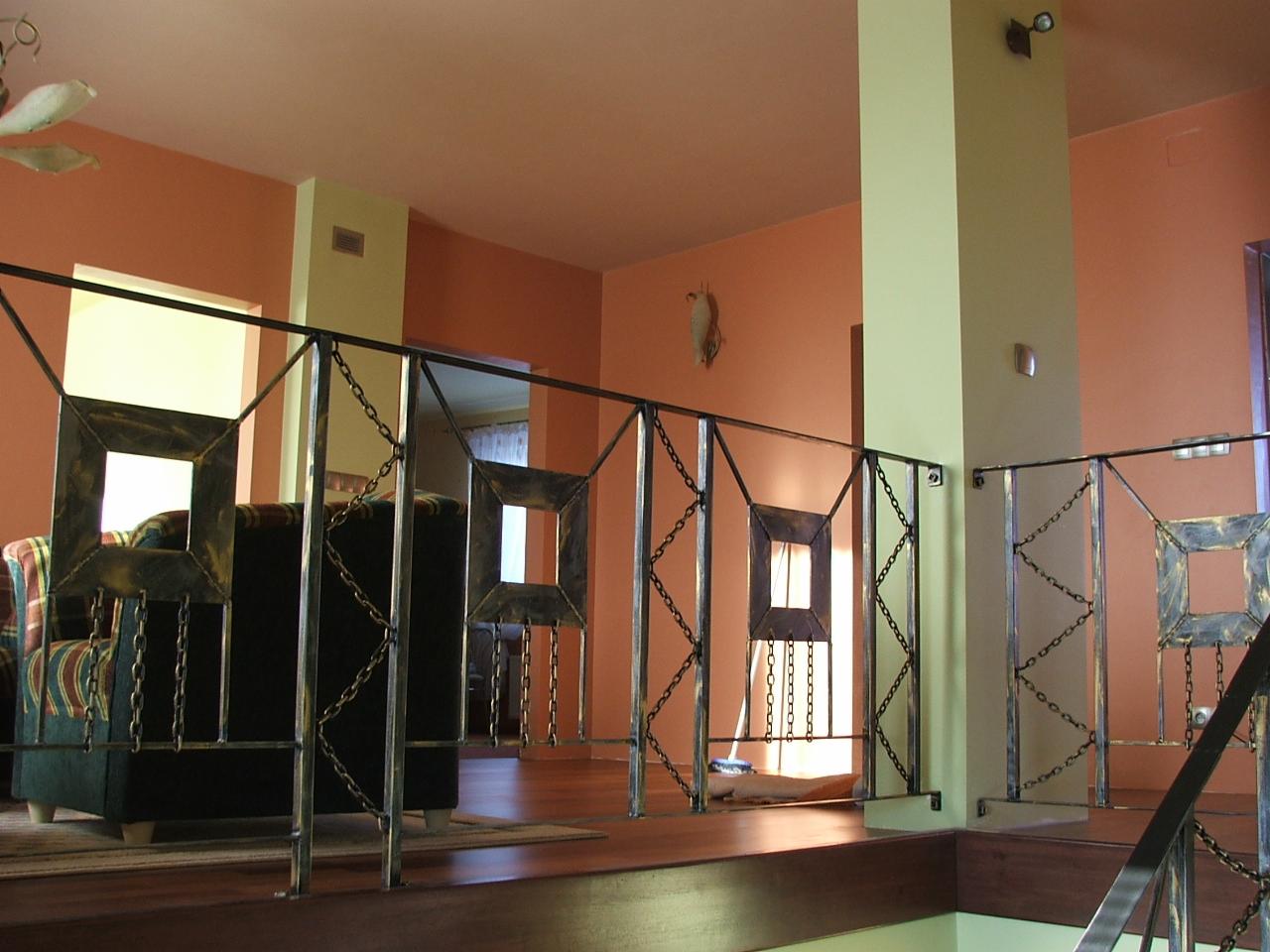 Nietypowa balustrada wewnętrzna z metalu, z kwadratowymi elementami dekoracyjnymi i łańcuchami, widok z góry na antresolę w pomarańczowym wnętrzu domu.
