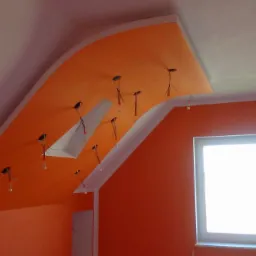 Pomarańczowe ściany i sufit z wykończeniem gipsowym i przygotowanymi punktami oświetleniowymi w poddaszowym pokoju z oknem.