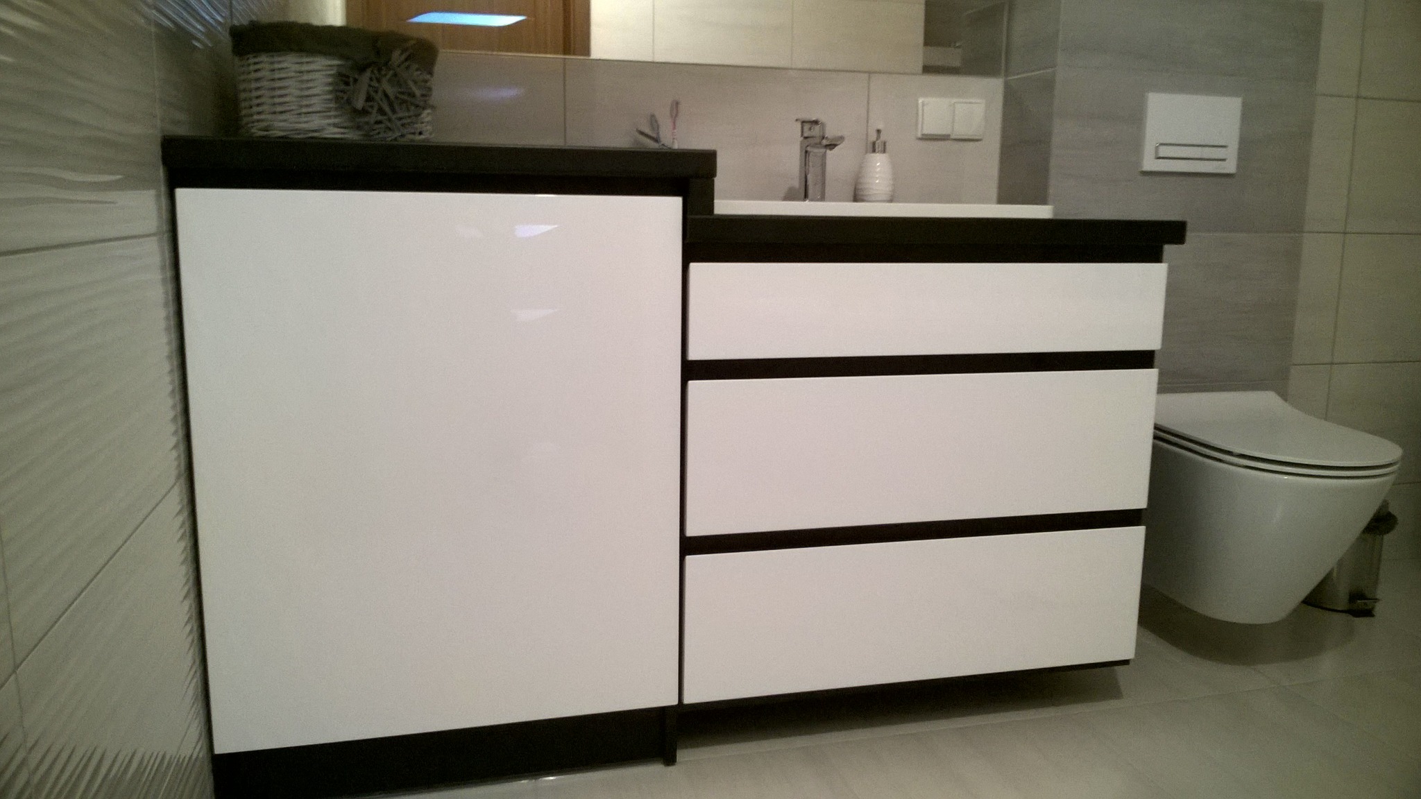 Biała szafka łazienkowa z czarnym blatem i trzema szufladami, obok toaleta podwieszana i koszyk dekoracyjny na blacie.