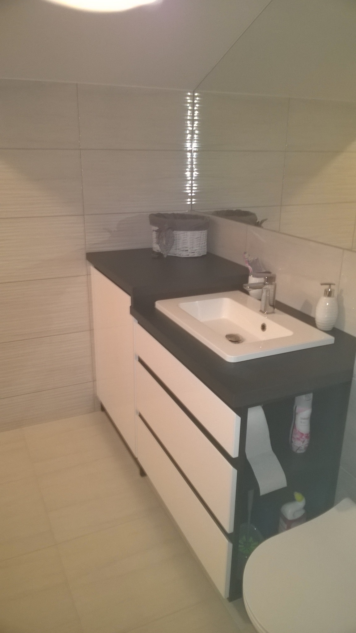 Nowoczesna umywalka nablatowa z czarnym blatem i białymi frontami szuflad, uzupełniona o wiklinowy koszyk i dozownik mydła na tle jasnych płytek.