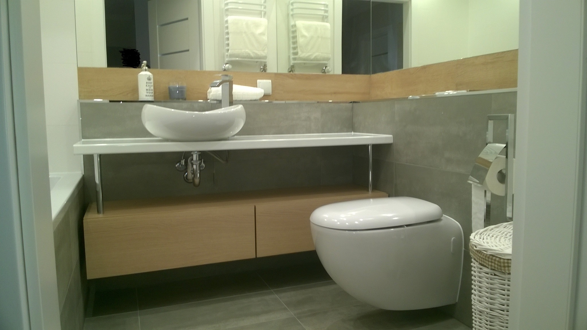 Nowoczesna łazienka z wiszącą toaletą, umywalką nablatową na białym blacie wspartym na chromowanych nóżkach, szafką z jasnego drewna i szarą glazurą.