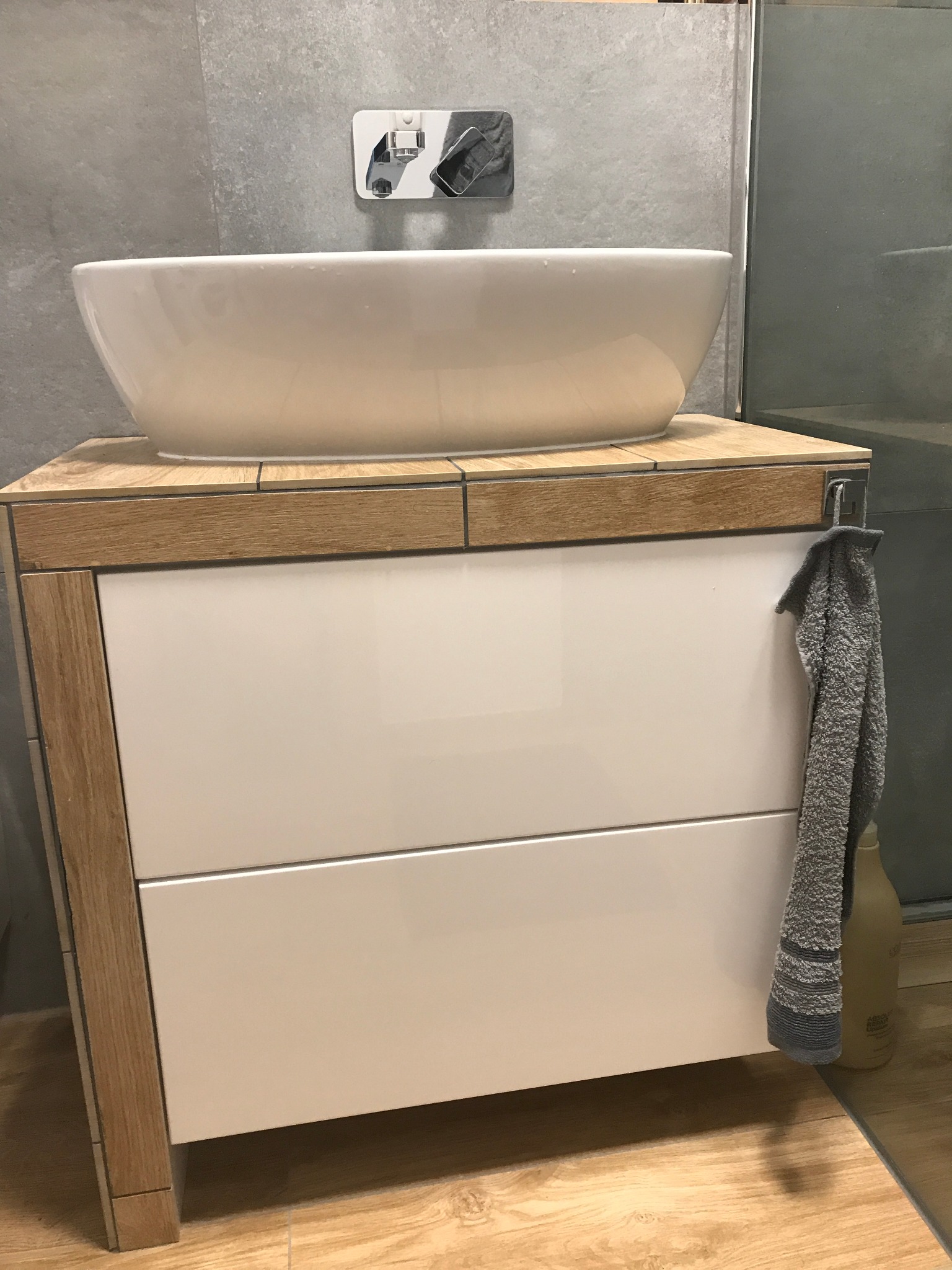 Nowoczesna łazienka z białą umywalką nablatową na szafce z drewnianymi akcentami, szary ręcznik wiszący z boku, w tle szara ściana i szklany parawan.