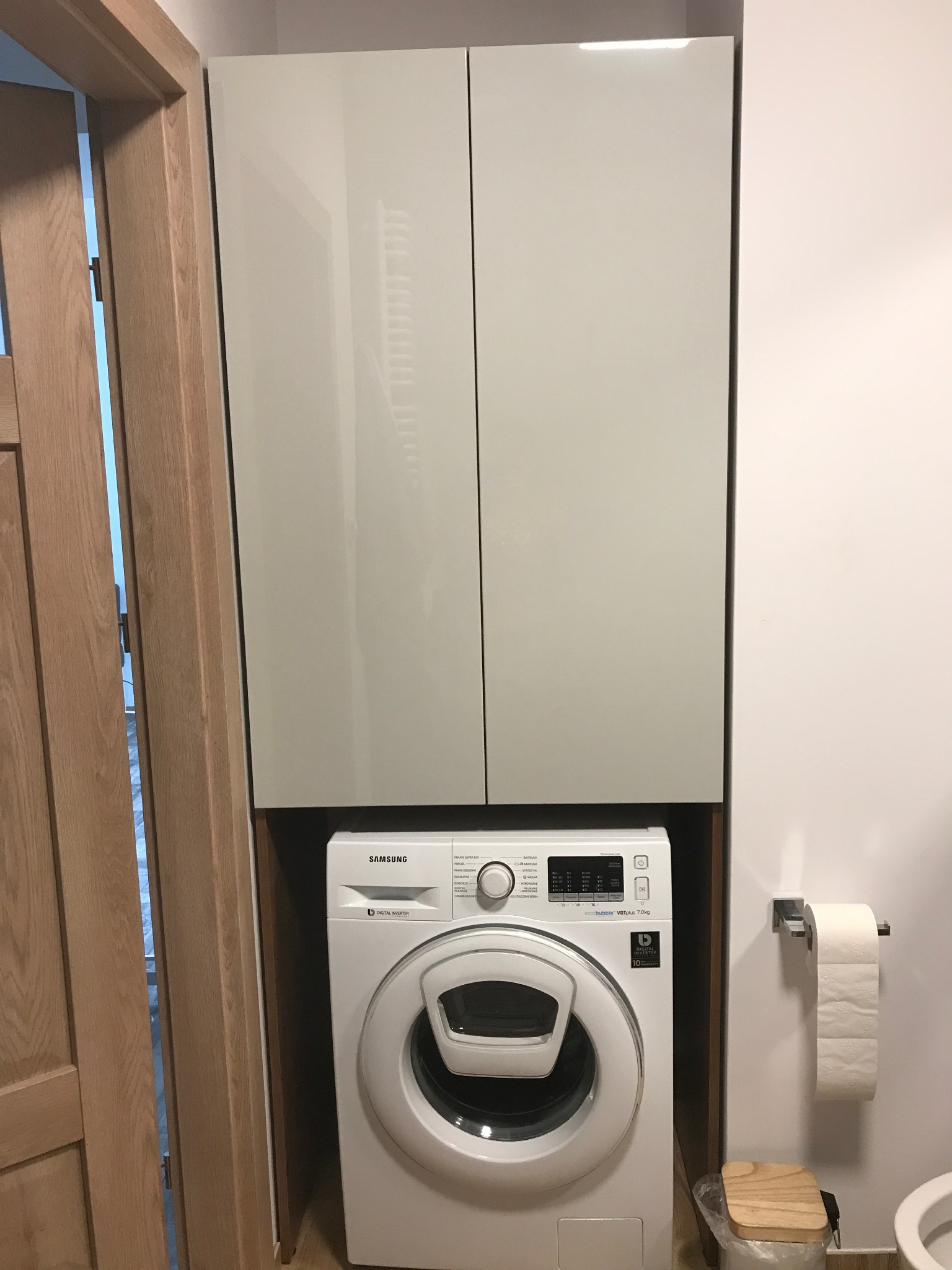 Zabudowa pralki w łazience z szafkami wiszącymi w kolorze jasnego beżu, widoczny fragment otwartych drzwi drewnianych po lewej stronie i toaleta z papierem toaletowym po prawej.