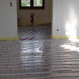 Układanie rur systemu grzewczego podłogowego na siatce montażowej w surowym stanie, widoczne taśmy maskujące wzdłuż ścian.