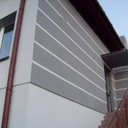 Elewacja budynku z szarymi pasami, widok z dołu, brązowa rynna i schody z barierką. Jasne tło nieba.