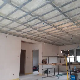Wnętrze w trakcie prac: metalowa konstrukcja sufitu podwieszanego z widoczną warstwą izolacji z pianki natryskowej, oświetlenie budowlane na trójnogu, rusztowanie z narzędziami, przygotowane ściany...